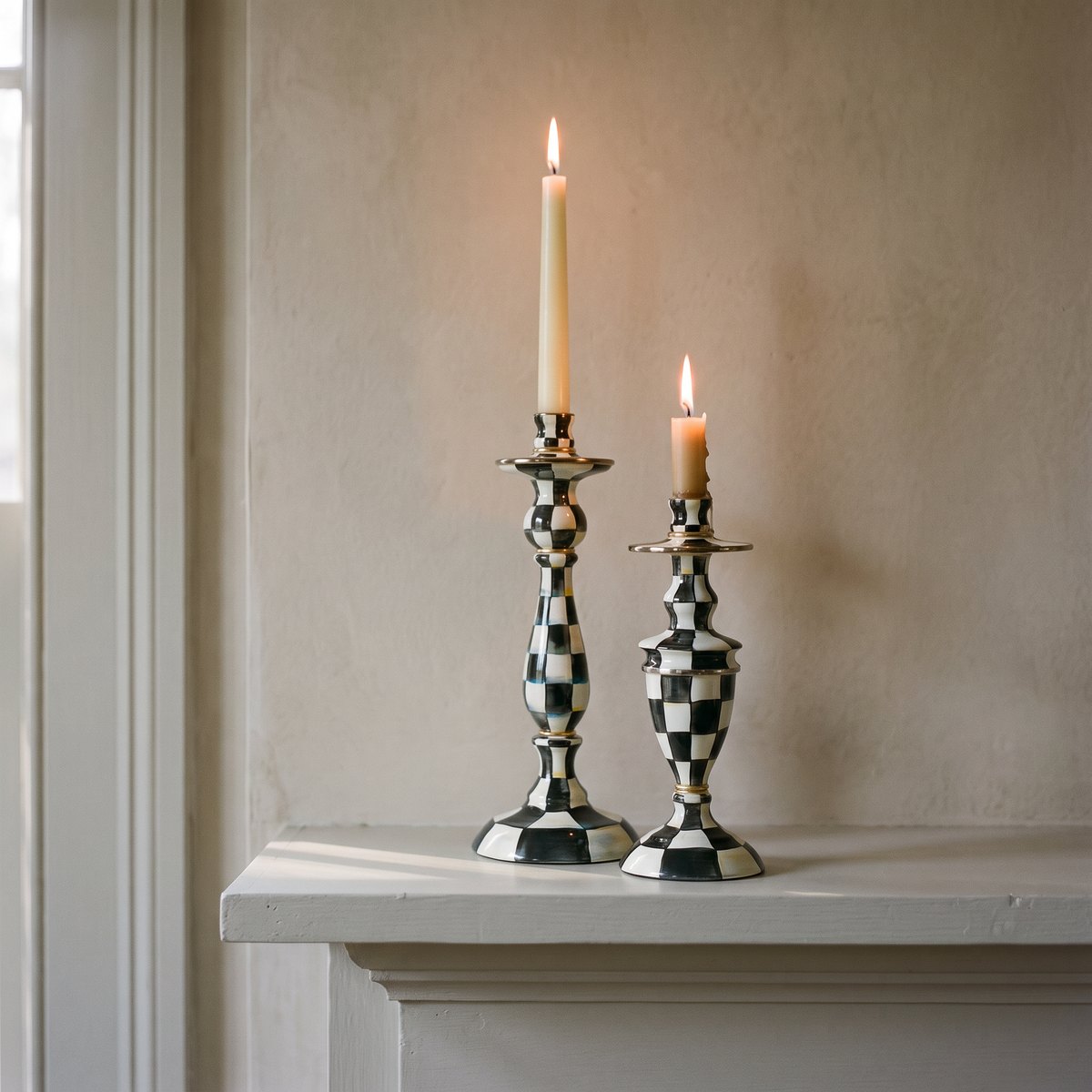 Large and medium MacKenzie-Childs Courtly Check enamel candlesticks holding lit cream tapers at one end of a mantel shelf