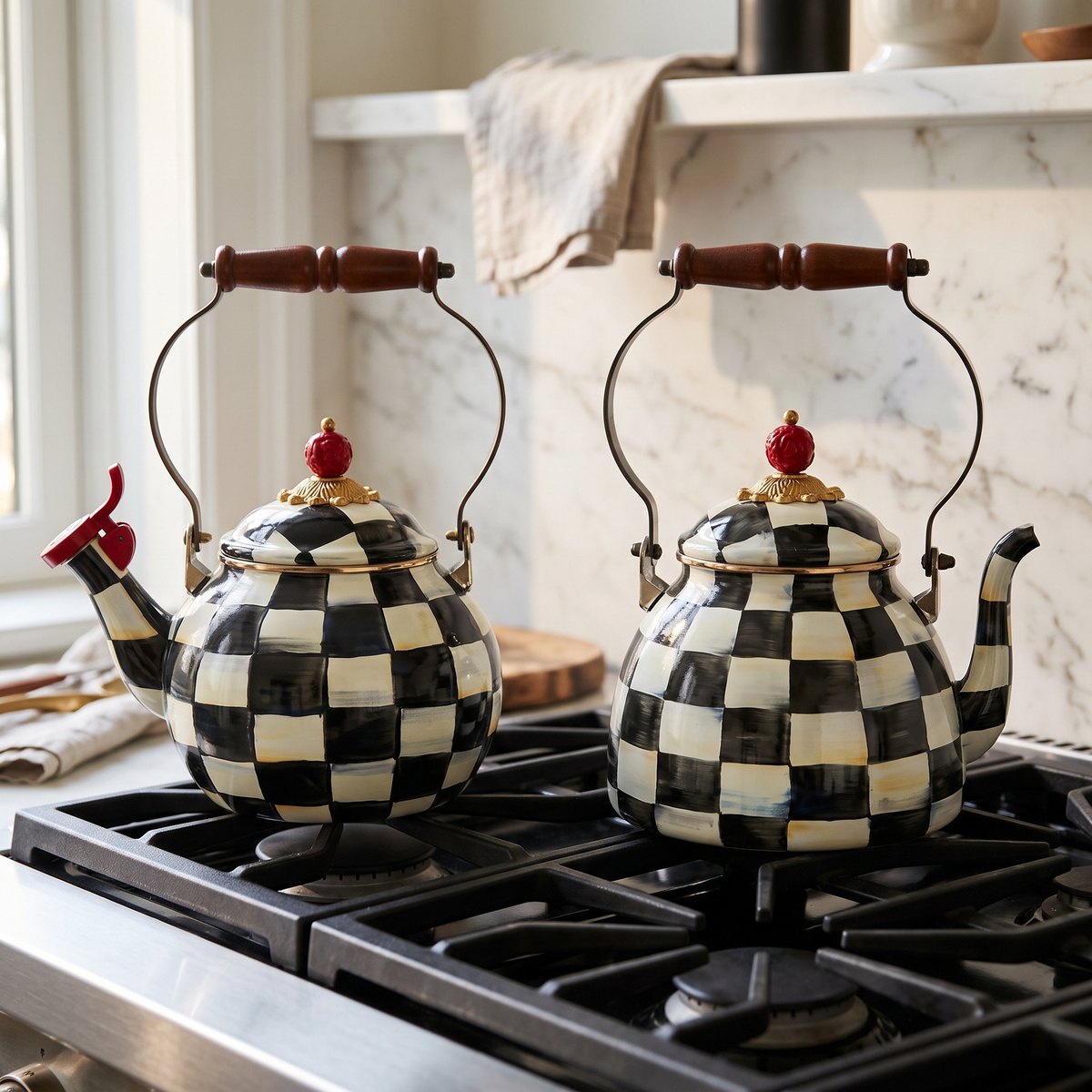 Two black and white MacKenzie-Childs Courtly Check enamel tea kettles side by side on a gas cooktop, the left one taller on a narrow base with a red whistle cap on its spout, the right one lower and sitting on a noticeably wider base