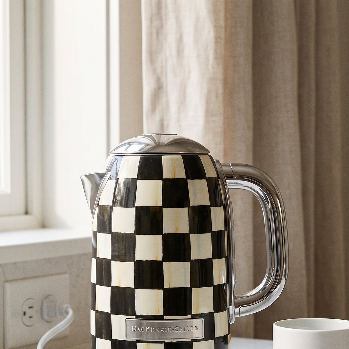 MacKenzie-Childs Courtly Check Electric Kettle on a white kitchen counter beside a window, a tall cylindrical black and white check body with a polished chrome handle and spout, with its cord running to a wall outlet