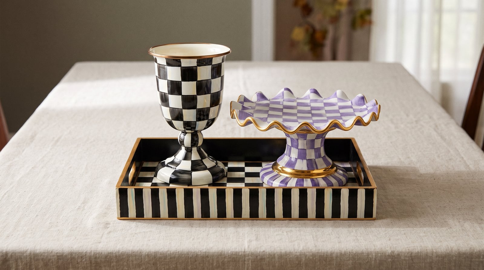 A MacKenzie-Childs Courtly Check pedestal vase and a Plum Check fluted cake stand grouped on a Courtly Check serving tray on a linen-covered fall dining table.