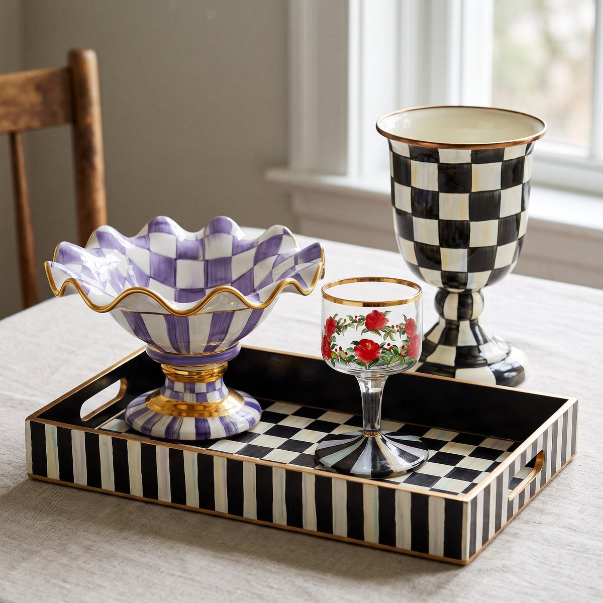 A MacKenzie-Childs Courtly Check serving tray laid flat, a Plum Check compote and a small hand-painted glass at mid height, and a Courtly Check pedestal vase standing tallest, showing three working heights on a fall table.