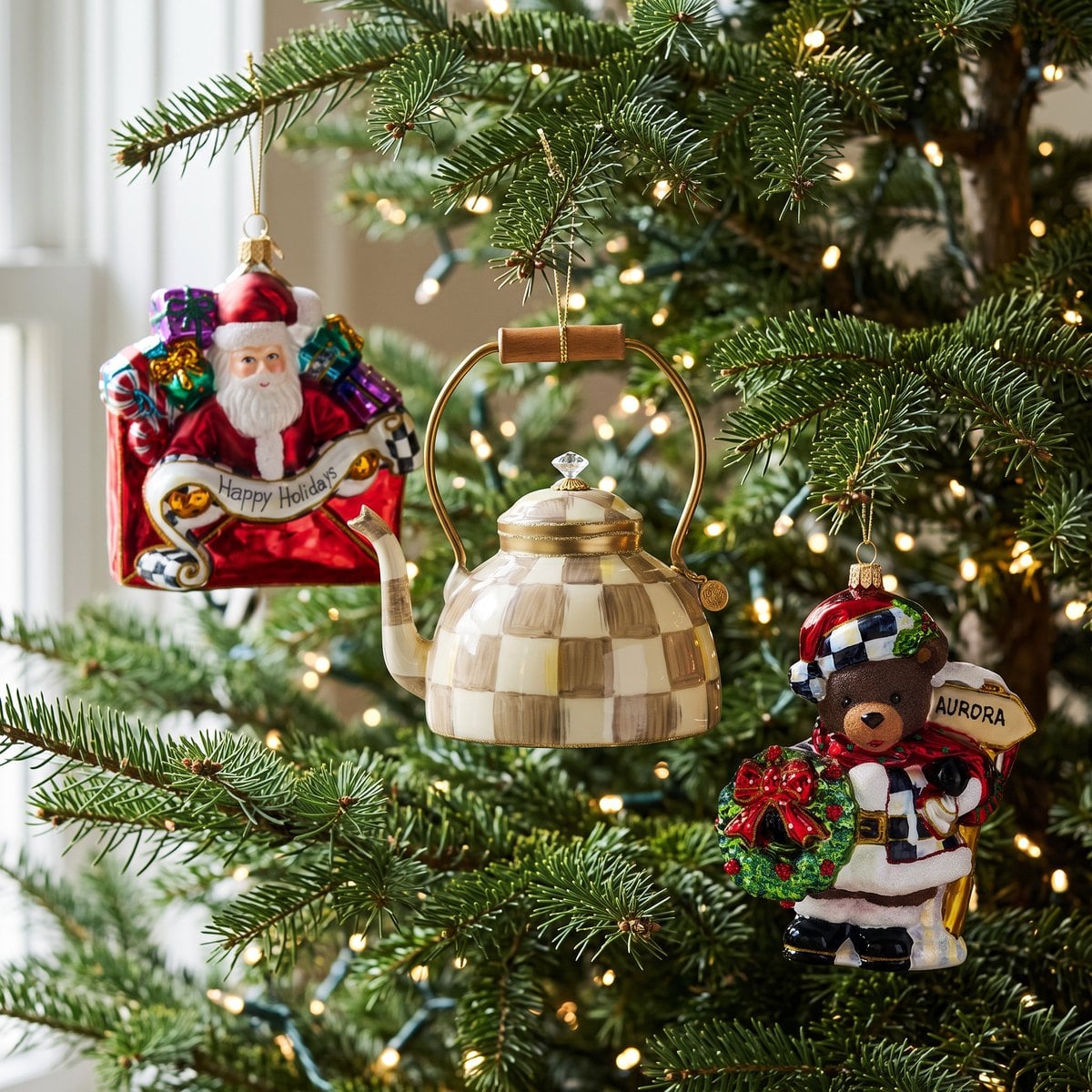 Close view of the Christmas Letter Santa, Mocha Check Tea Kettle and Aurora Teddy Bear glass ornaments hanging at different heights among fir branches