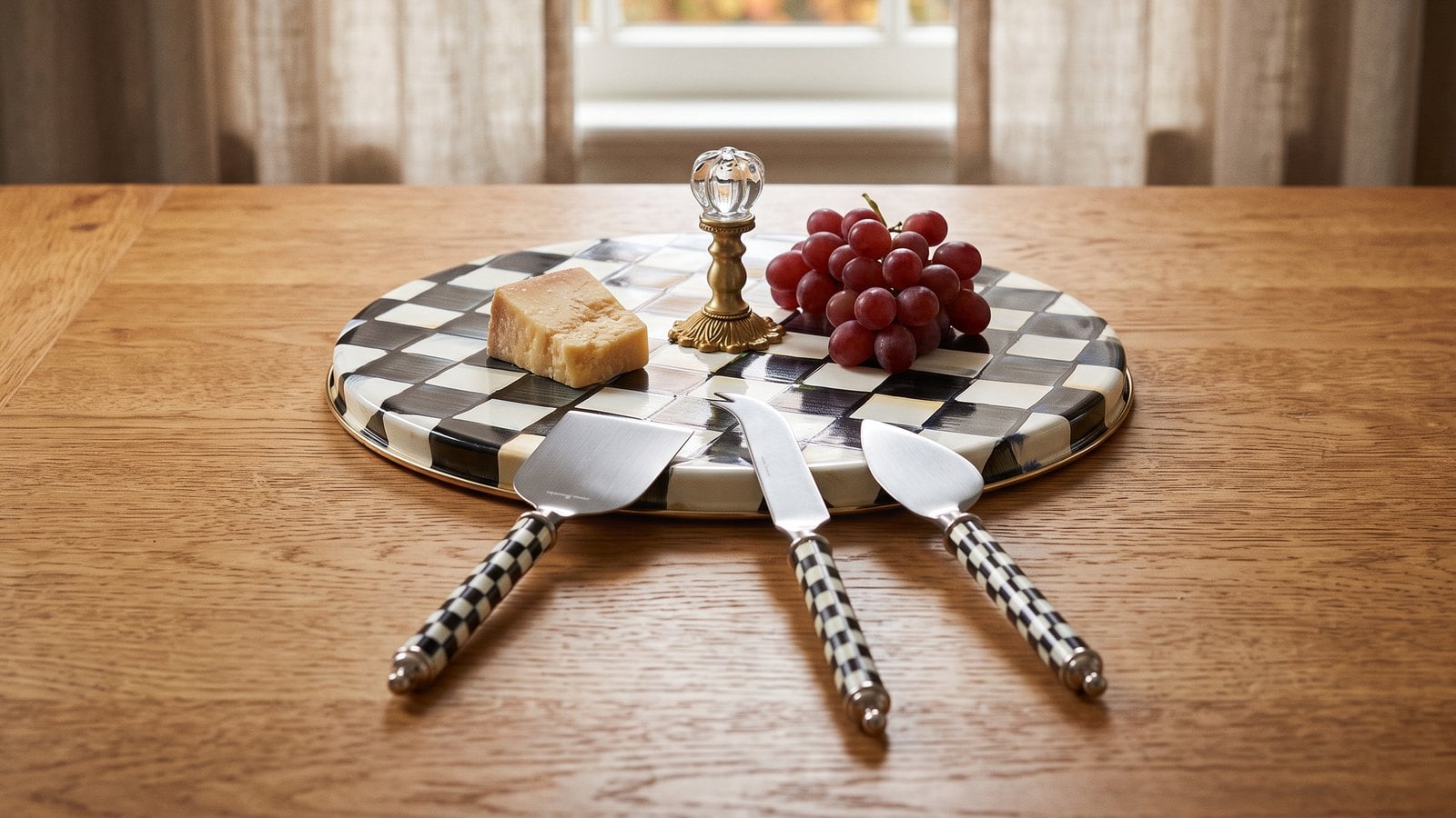 Courtly Check enamel cheese course with a brass and glass knob handle, holding cheese and red grapes, with three Courtly Check cheese knives laid in front on an oak dining table